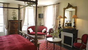 Een hemelbed, een rode fauteuil, een tafeltje met een vaas, een open haard en een grote spiegel.