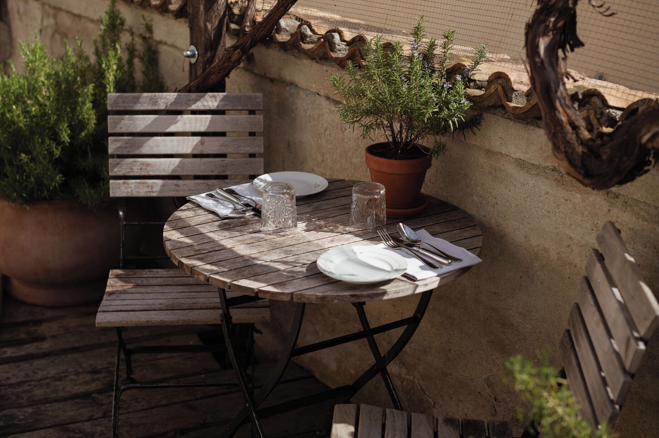 A wooden table set with plates, cutlery, and glasses, surrounded by benches and a potted plant.