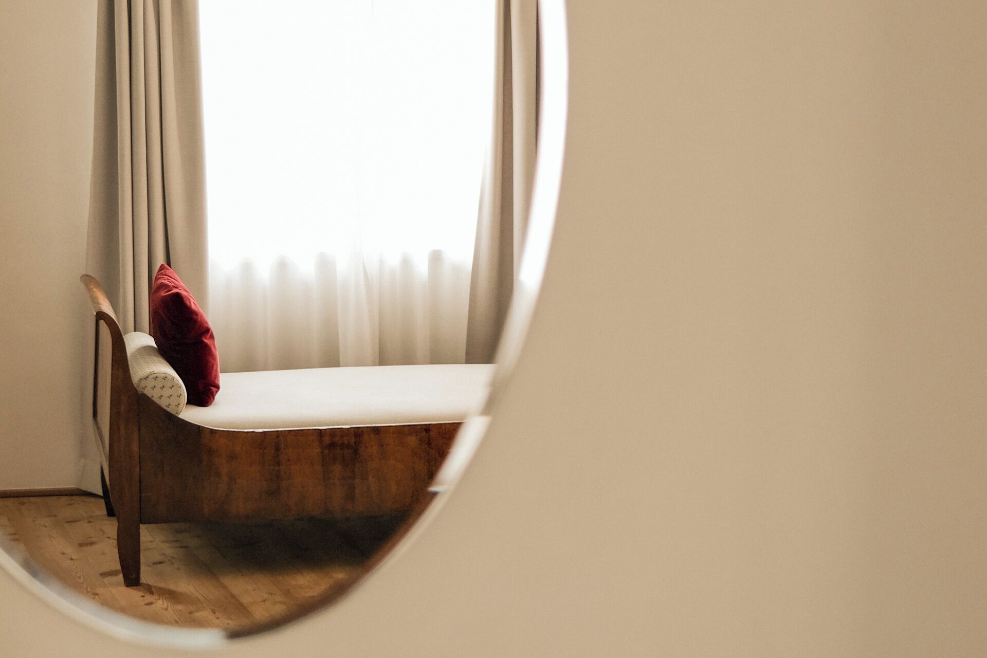 Un espejo redondo que refleja una esquina de una habitación con una silla de madera y un cojín rojo.