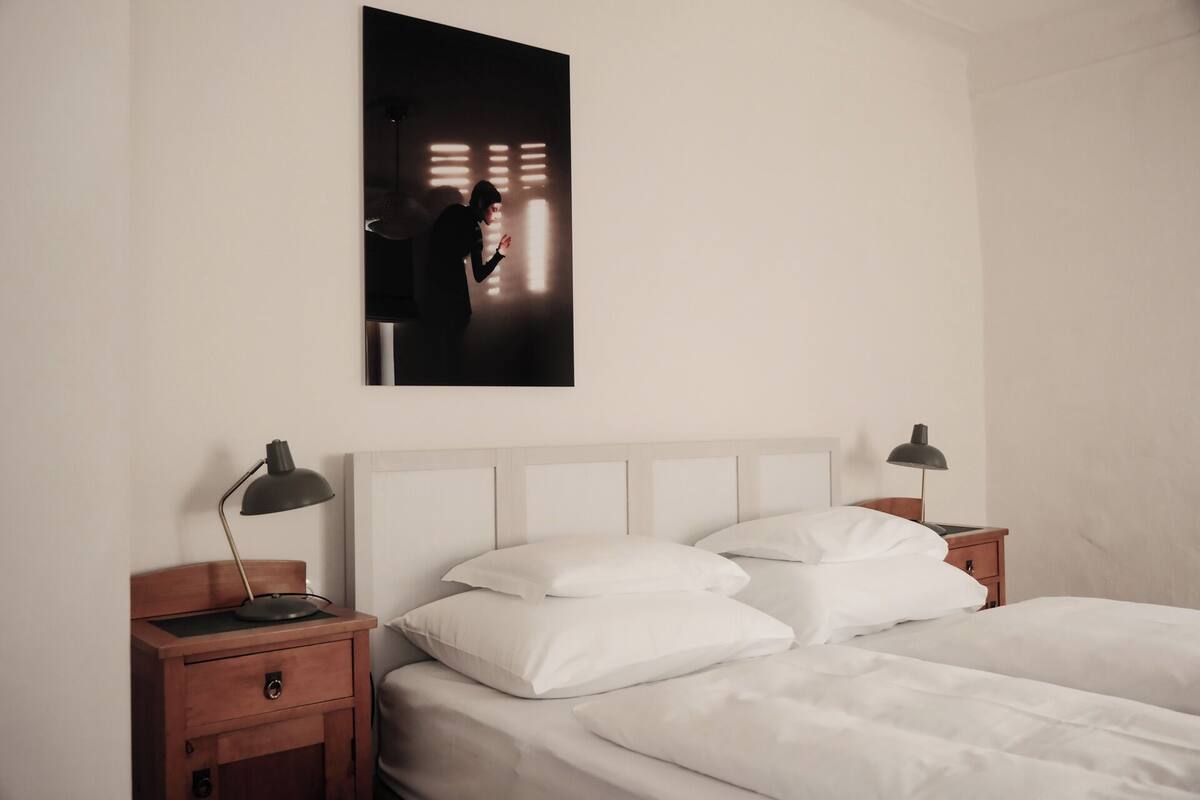 A bedroom with a bed, two bedside tables, a nightstand, a wall-mounted picture, and a ceiling light fixture.