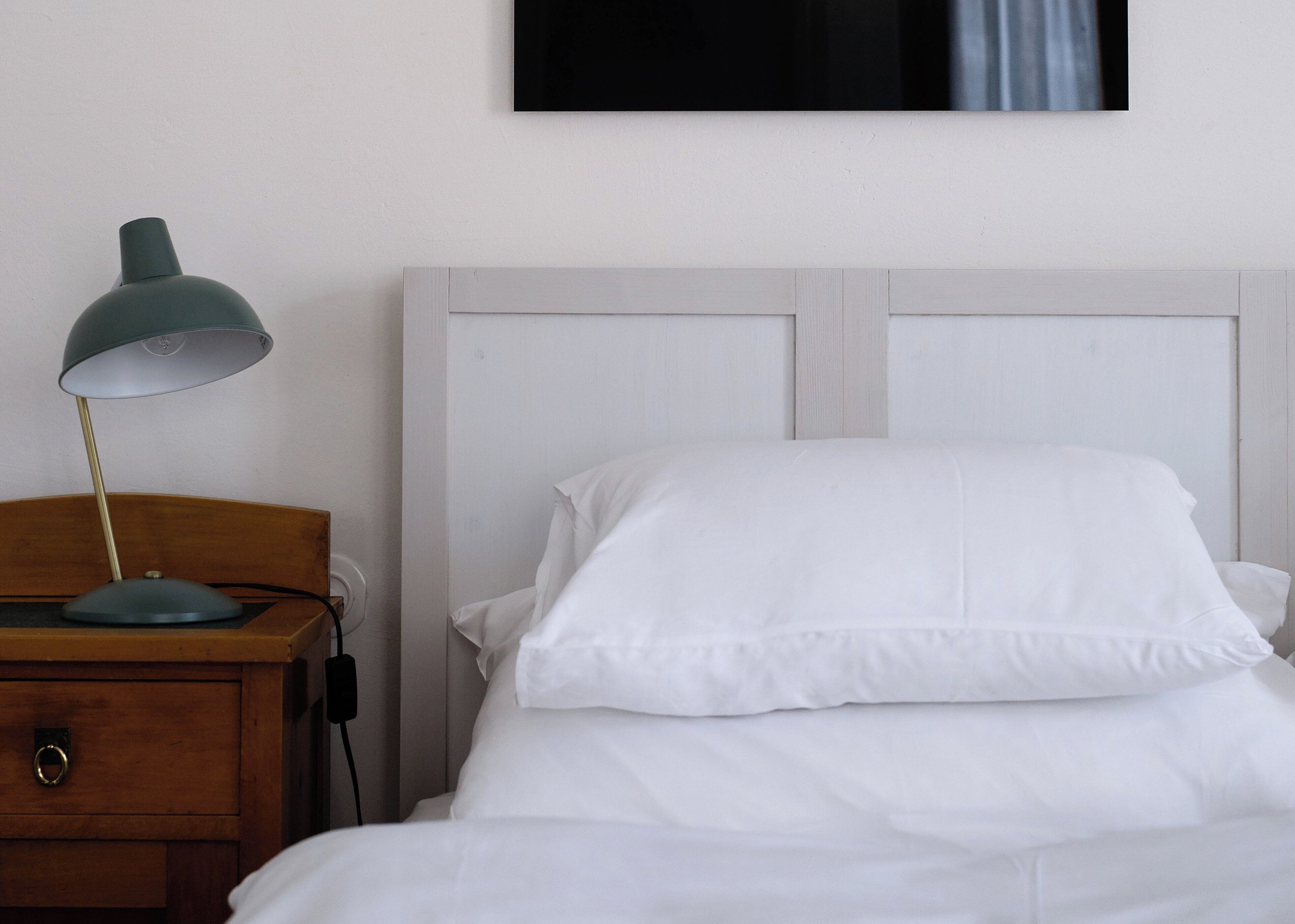 A neatly made bed with white linens, a wooden nightstand with a green desk lamp, and a wall-mounted TV above the bed.
