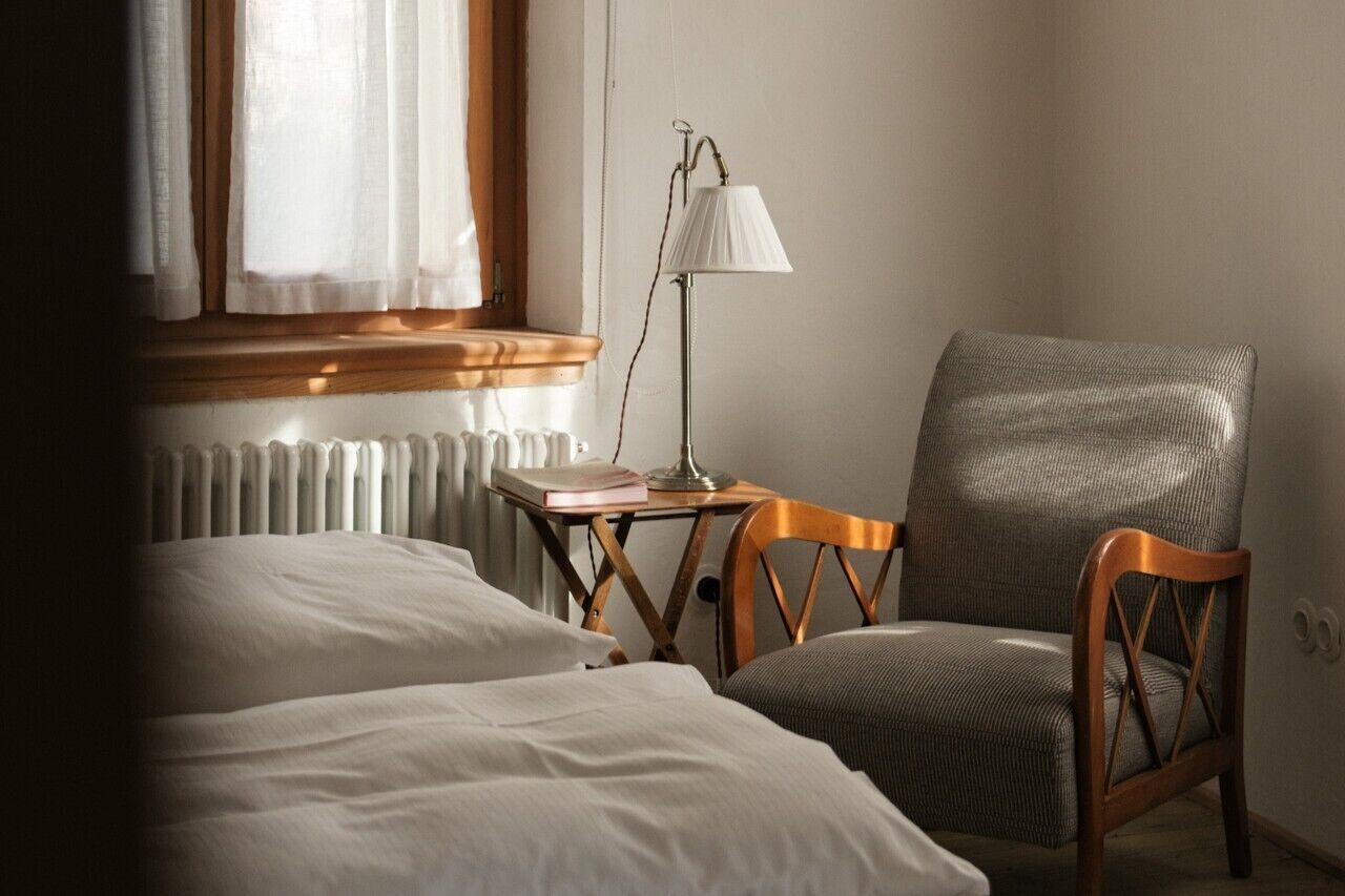 Hotellihuone, jossa on kaksi sänkyä, puinen tuoli, pieni pöytä lampun kanssa ja ikkuna, jossa on verhot.