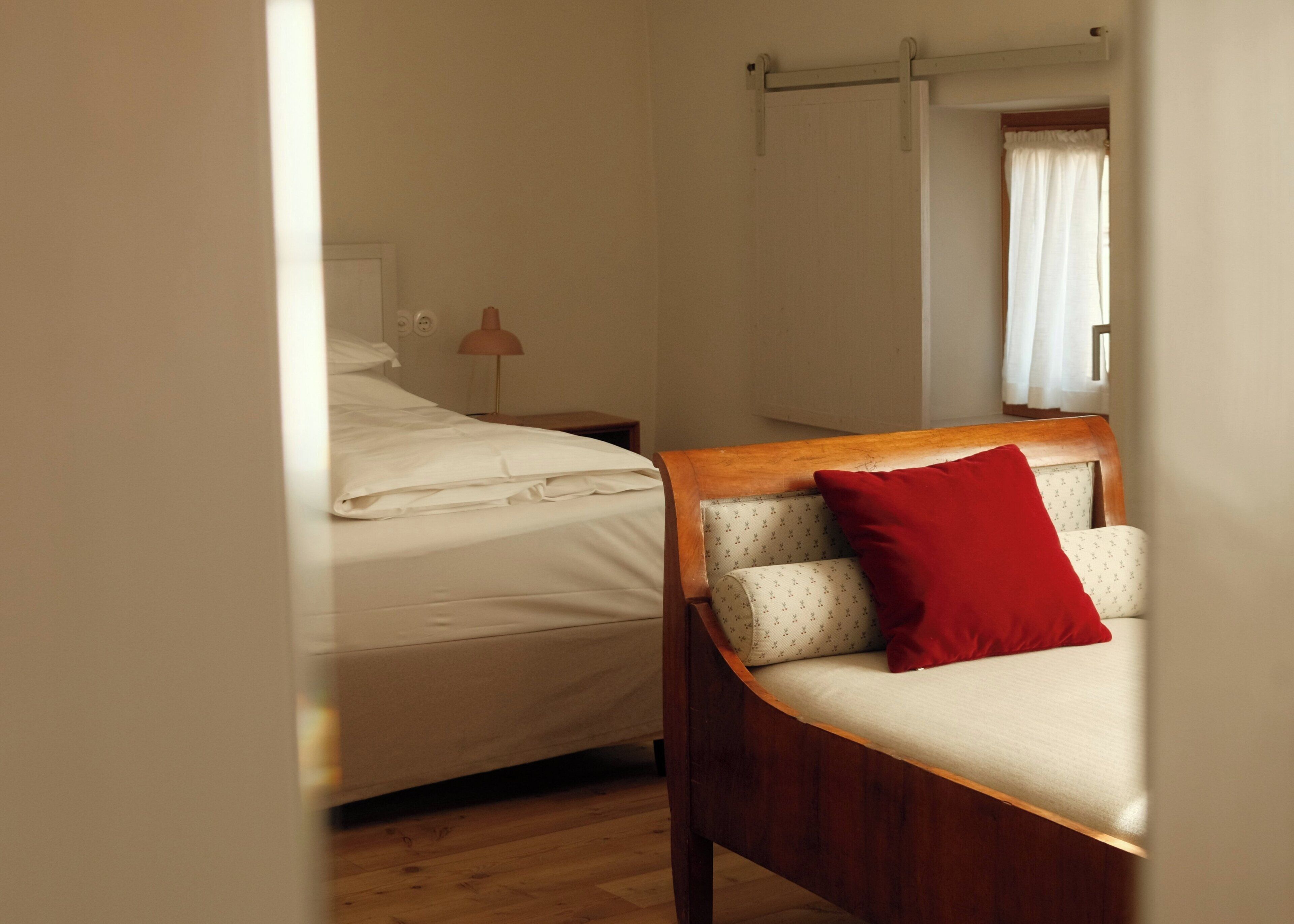 Une chambre avec un cadre de lit en bois, un matelas blanc et un oreiller rouge. On y trouve une table de chevet avec une lampe et une fenêtre avec des rideaux.