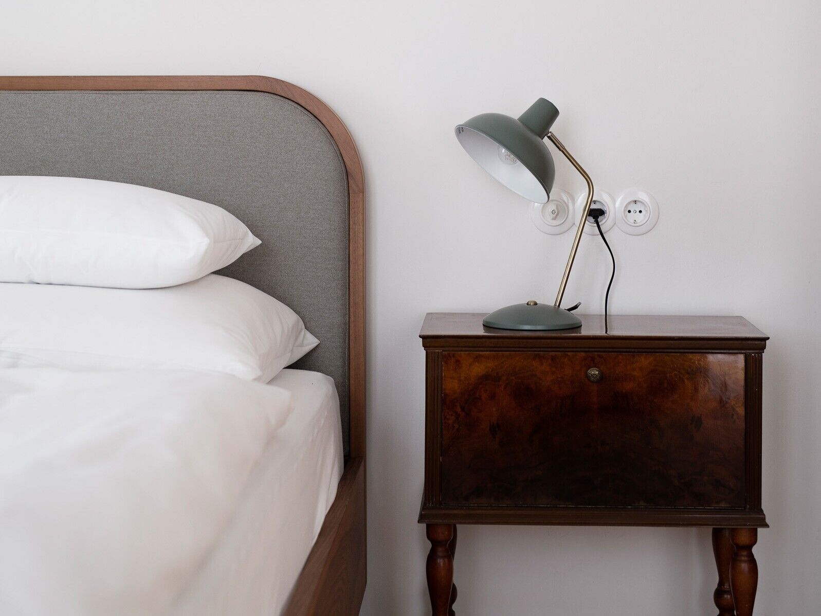 En seng med hvide sengetøj og puder, et trænatbord med en lampe og en stikkontakt.