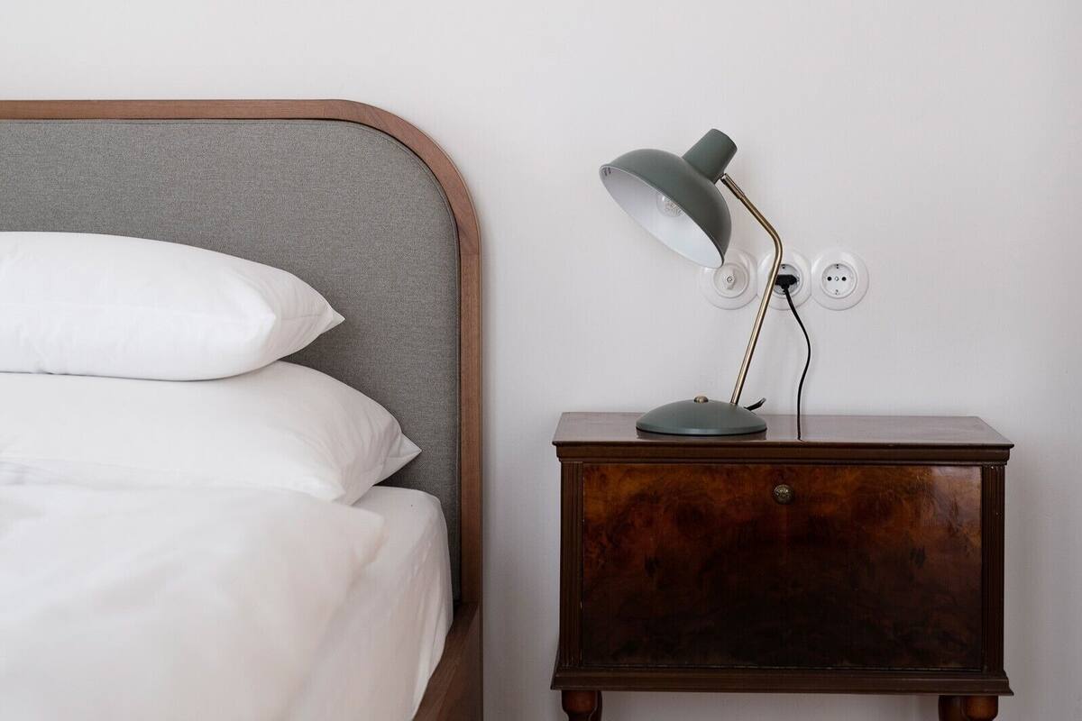 A bed with white linens and pillows, a wooden nightstand with a lamp, and a wall socket.