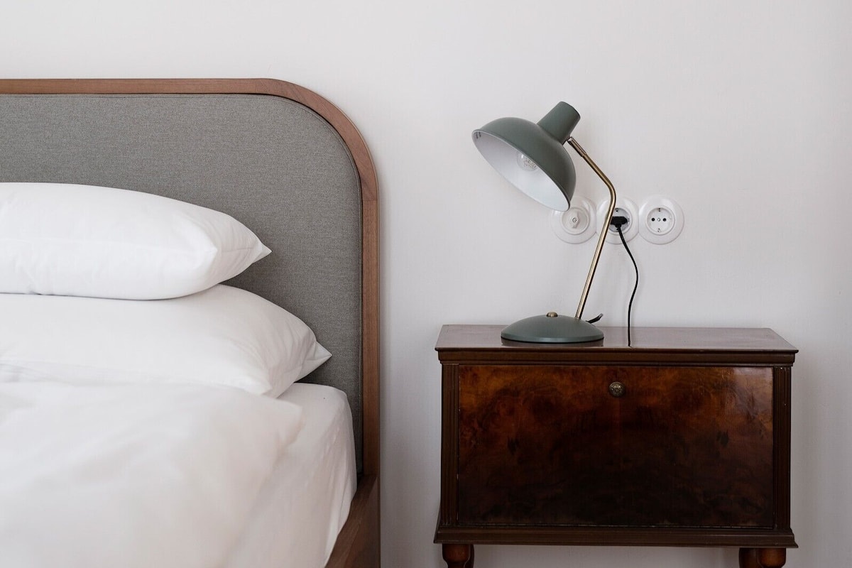 A bed with white linens and pillows, a wooden nightstand with a lamp, and a wall socket.