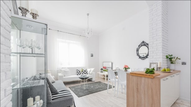 Una sala de estar moderna con un sofá gris, una vitrina de cristal con copas de vino, una isla de madera con tabla de cortar y una pared de ladrillo blanco decorativa.