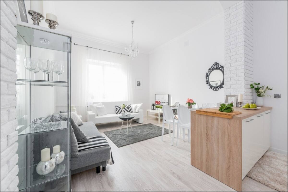 Una sala de estar moderna con un sofá gris, una vitrina de cristal con copas de vino, una isla de madera con tabla de cortar y una pared de ladrillo blanco decorativa.
