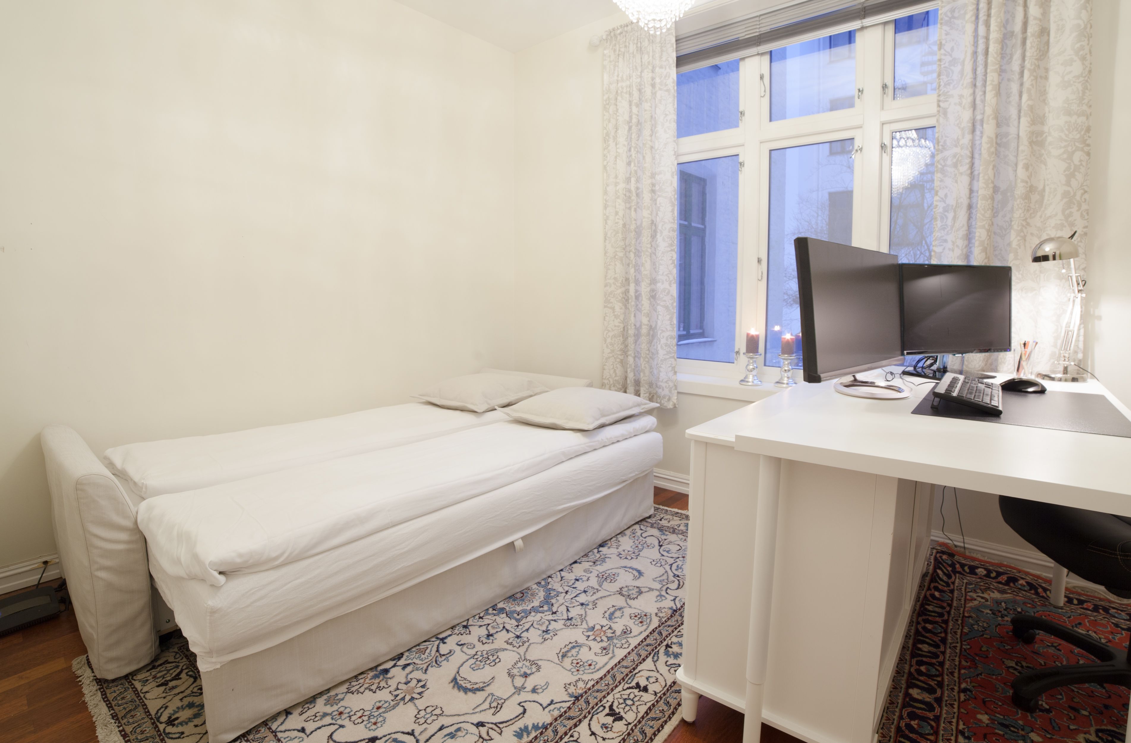 A small, neatly arranged room with a single bed, a desk with a computer, and a window with curtains.
