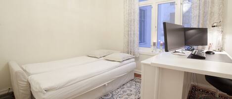 A small, neatly arranged room with a single bed, a desk with a computer, and a window with curtains.