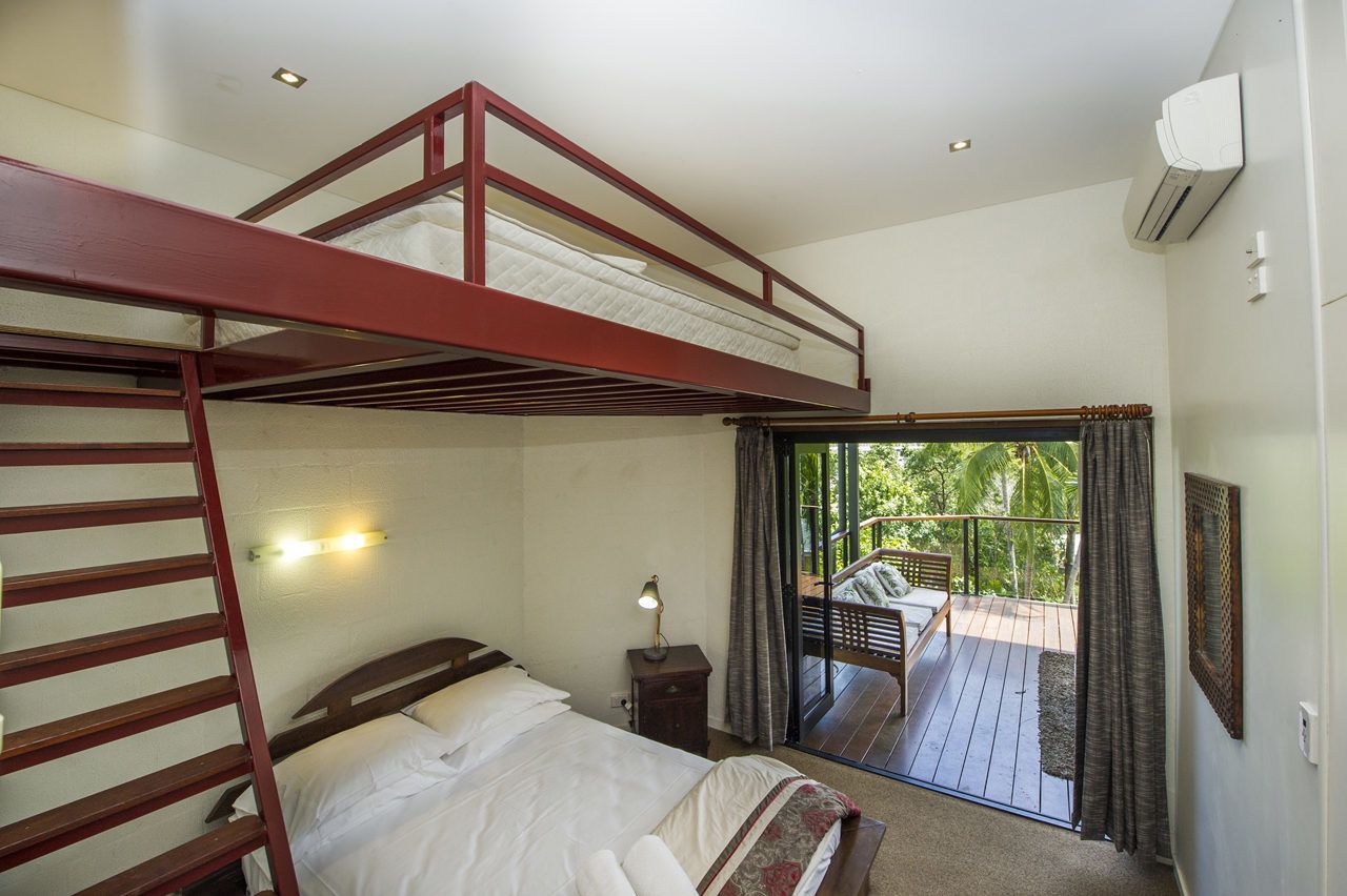 A bedroom with a bunk bed, a bedside table with a lamp, a window with curtains, and a balcony with a chair.
