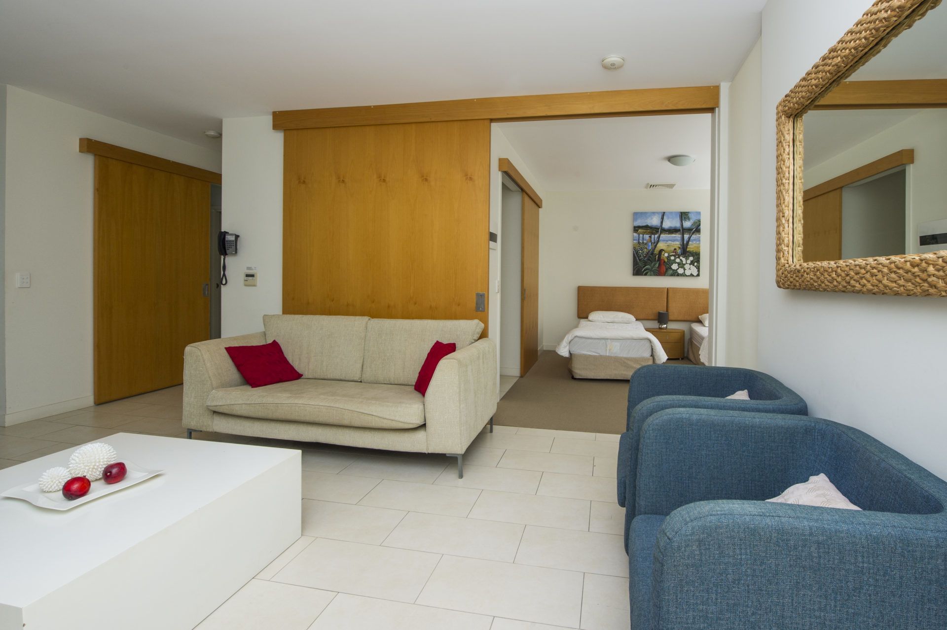 Een moderne hotelkamer met een beige bank, een blauwe bank, een witte salontafel en een bed met een houten hoofdeinde.