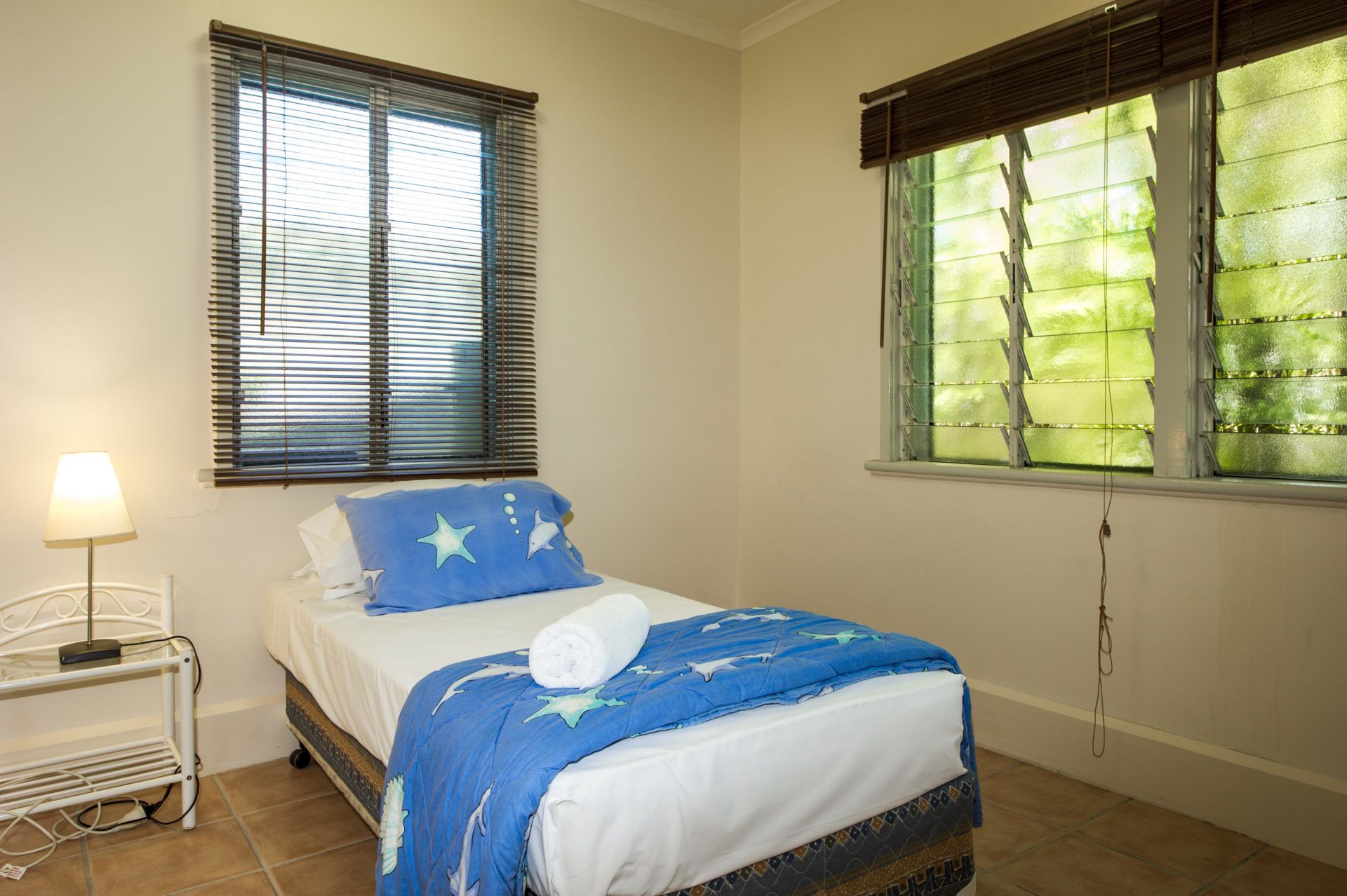 A bedroom with a bed, a nightstand, a lamp, and two windows with blinds.