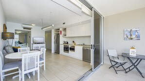 Una cocina moderna con armarios blancos, horno empotrado y refrigerador. Hay una zona de comedor con sillas blancas y una mesa.