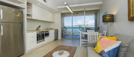 A modern kitchen with a stainless steel refrigerator, white cabinets, and a wooden dining table. There is a balcony with a view of the sea.