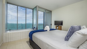 Un dormitorio con un ventanal que ofrece vistas al mar, una cama con ropa blanca y un televisor sobre un soporte de madera.