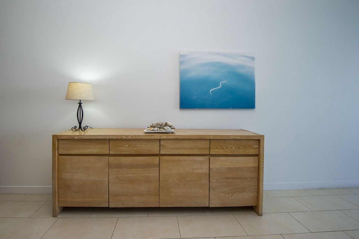 A wooden cabinet with drawers and a lamp on top, against a plain wall with a blue painting.