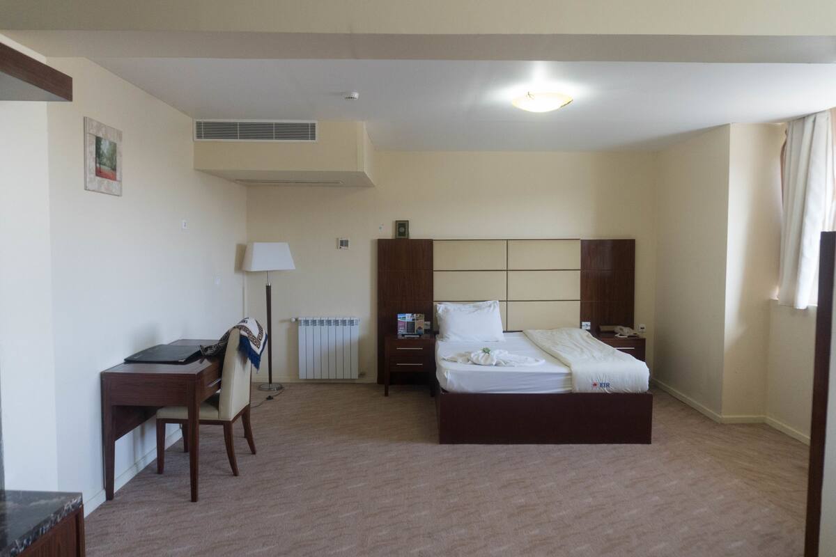 A hotel room with a bed, a desk, a chair, a nightstand, a lamp, and a radiator.