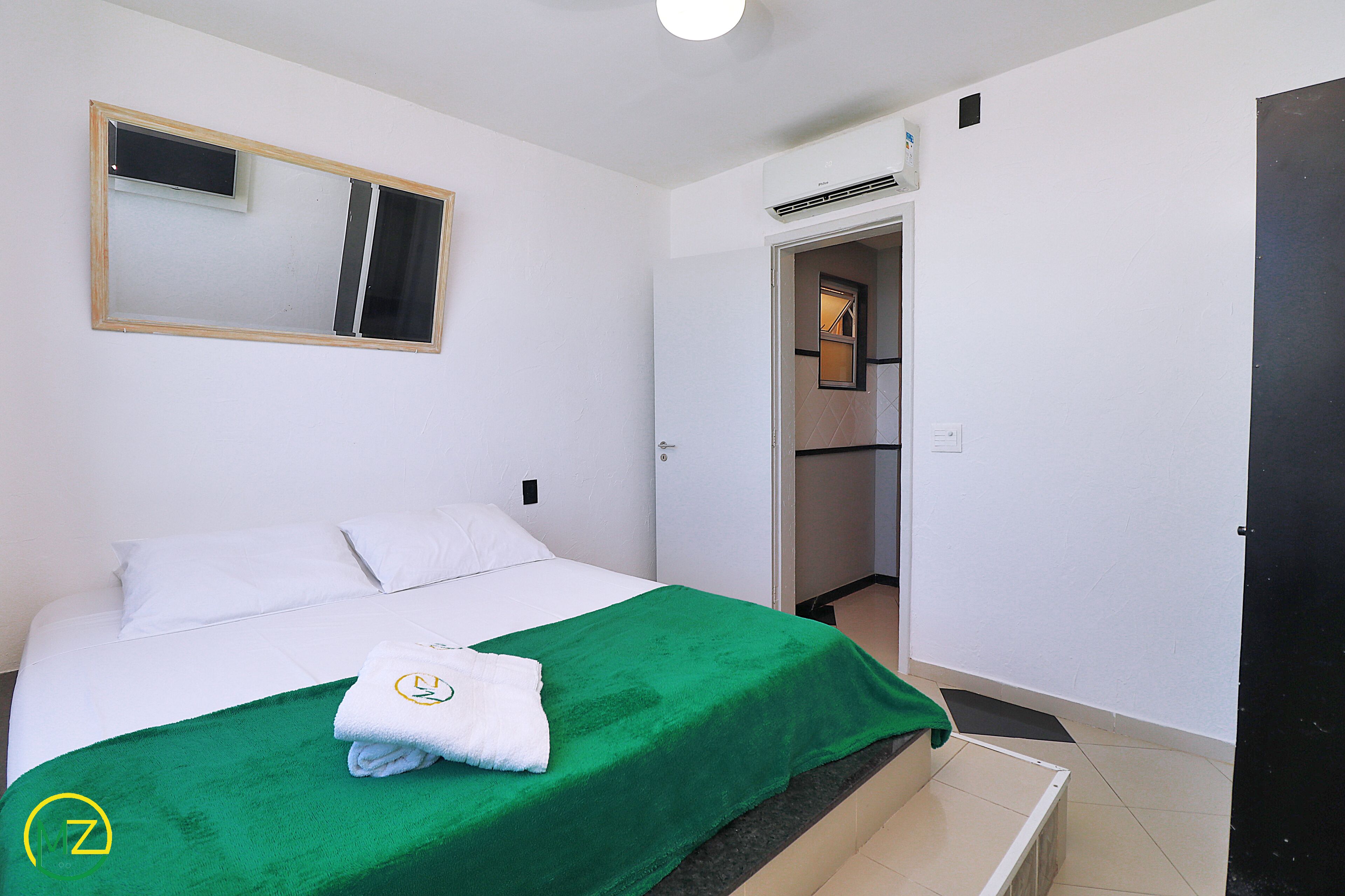 Habitación de hotel con cama, manta verde, almohada blanca, toalla, espejo y puerta que da a un baño.