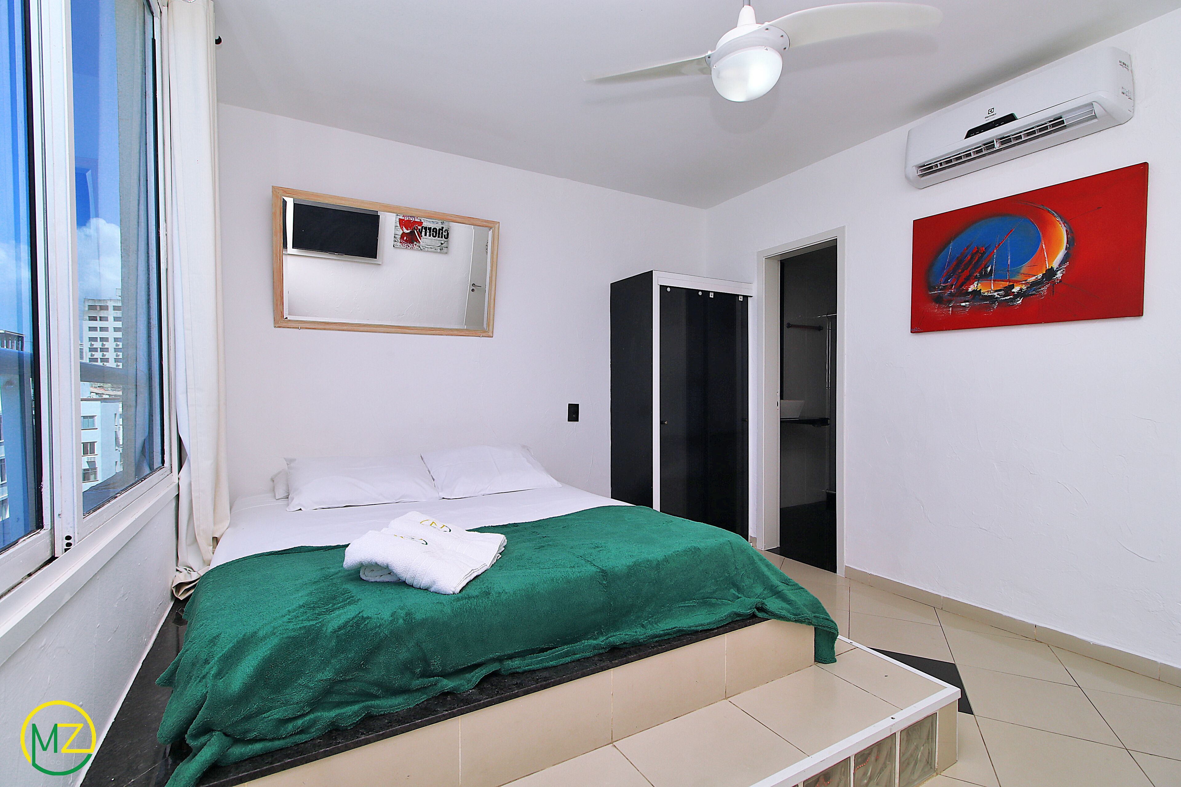Un dormitorio con una cama, un edredón verde, una almohada blanca, un ventilador de techo, un aire acondicionado, un cuadro enmarcado y una obra de arte roja.