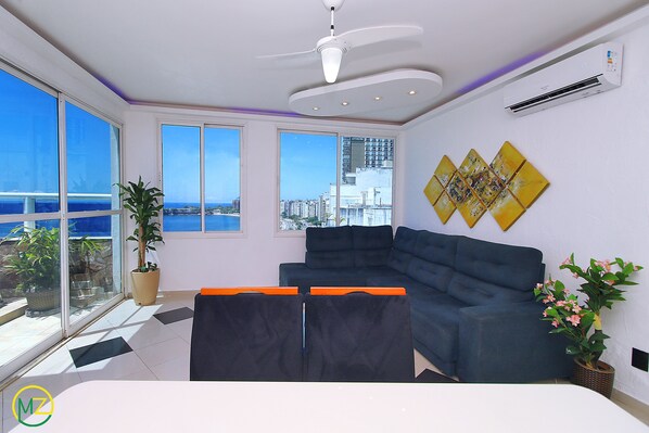 A modern living room with a large sofa, a ceiling fan, and a view of the city and ocean through large windows.