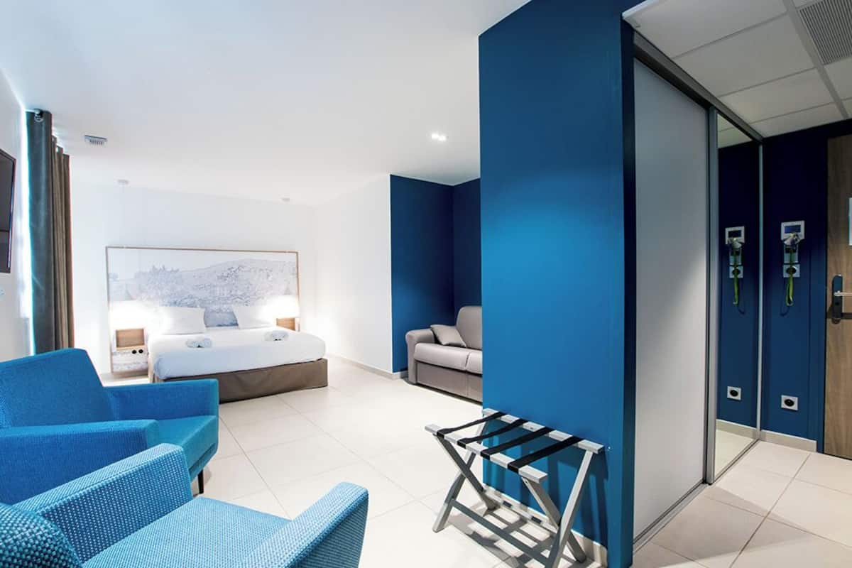 Ein modernes Hotelzimmer mit einem Bett, einem blauen Sofa, einem kleinen Tisch und einer Schiebetür zum Badezimmer.