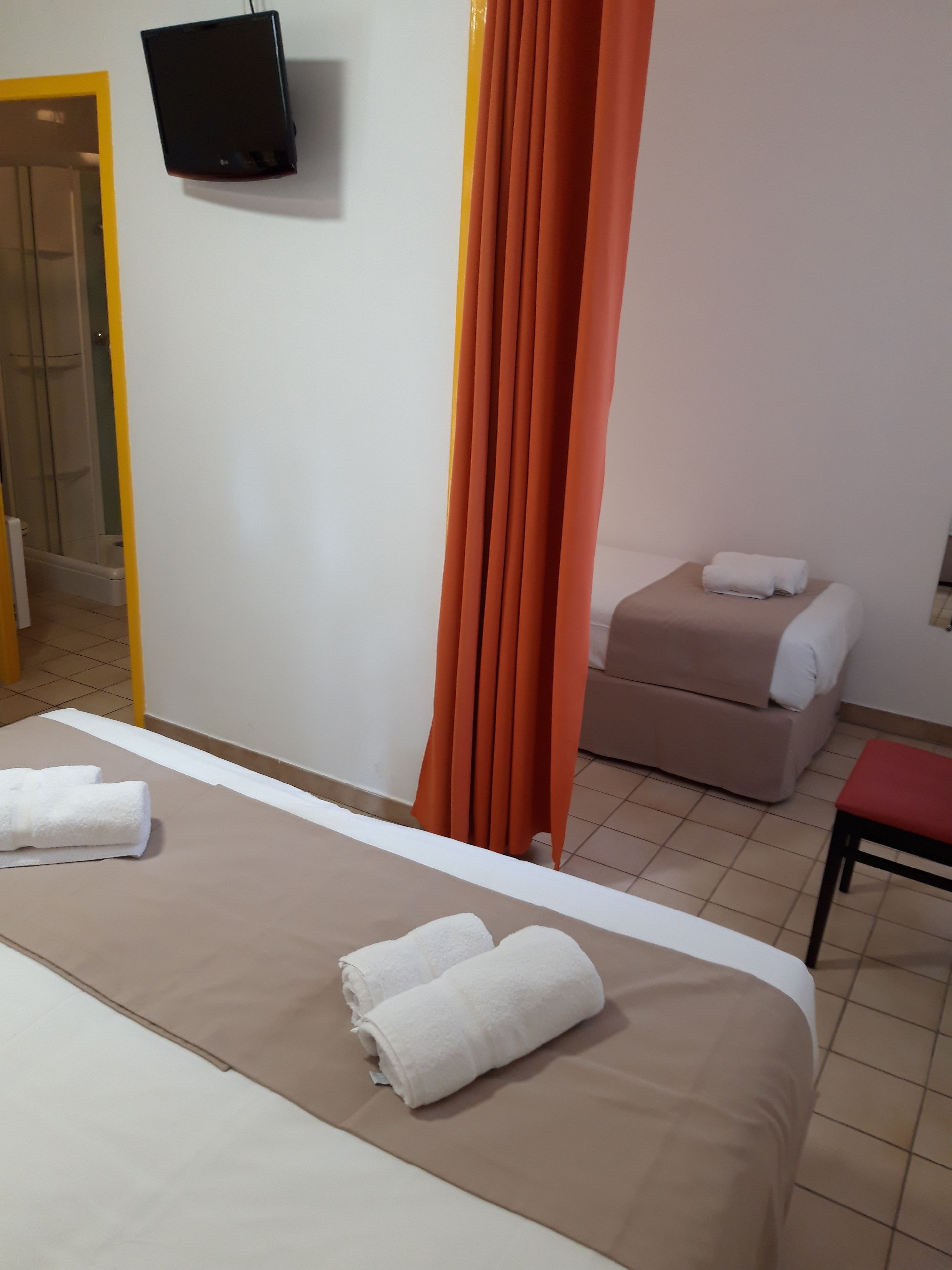Une chambre d’hôtel avec un lit, un téléviseur fixé au mur, un espace douche et un petit coin salon avec un fauteuil rouge.