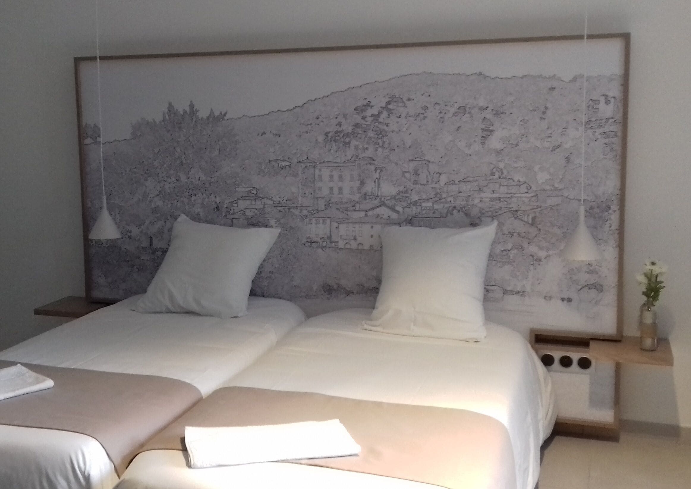 Una cama doble con dos almohadas blancas, una mesita de noche con un florero y un mural en la pared con vista a un paisaje urbano.