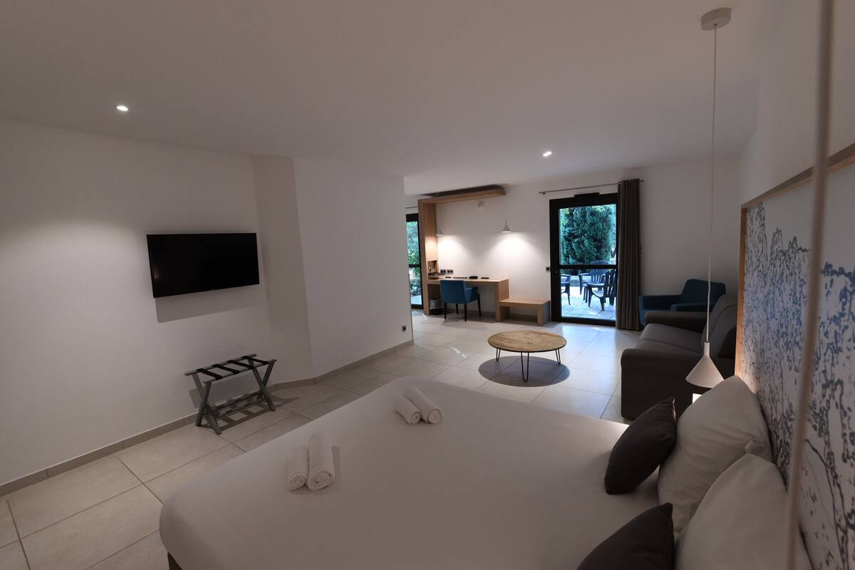 Ein modernes Hotelzimmer mit einem großen Bett, einem flachbildfernseher an der Wand, einem kleinen Tisch und einer Sitzecke mit Sofa und Sesseln.