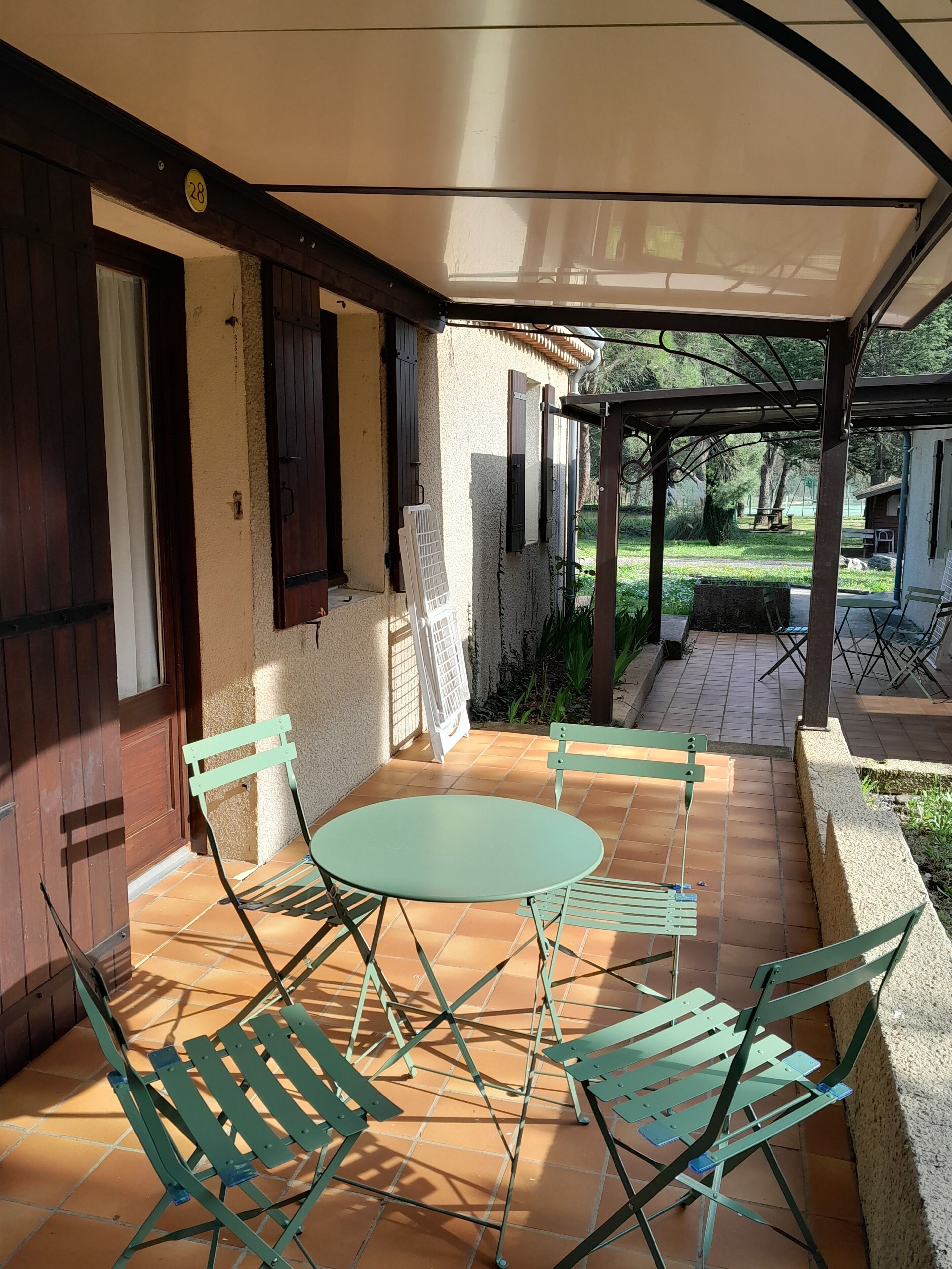 Un patio cubierto con una mesa redonda y cuatro sillas, una silla plegable blanca, y un pequeño jardín visible a través del lado abierto.