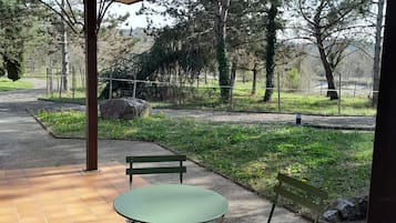 Un área de estar al aire libre, techada, con una mesa y dos sillas, rodeada por un jardín con árboles y un sendero.