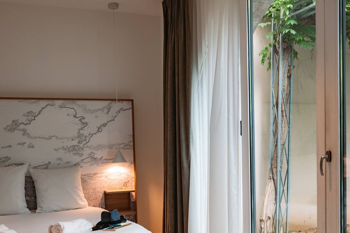 Ein Schlafzimmer mit Marmorkopfteil, einem Bett mit weißen Bettwäsche, einer Nachttischlampe und Blick durch eine Glastür auf einen Außenbereich mit Baum und Metallkonstruktion.