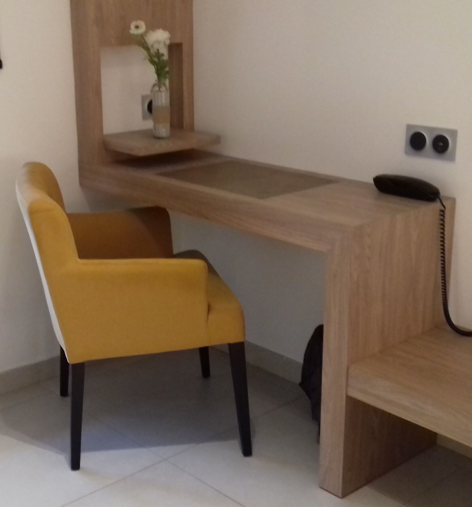 Un escritorio moderno con detalles de madera, una silla amarilla y un pequeño jarrón con flores sobre una estantería.