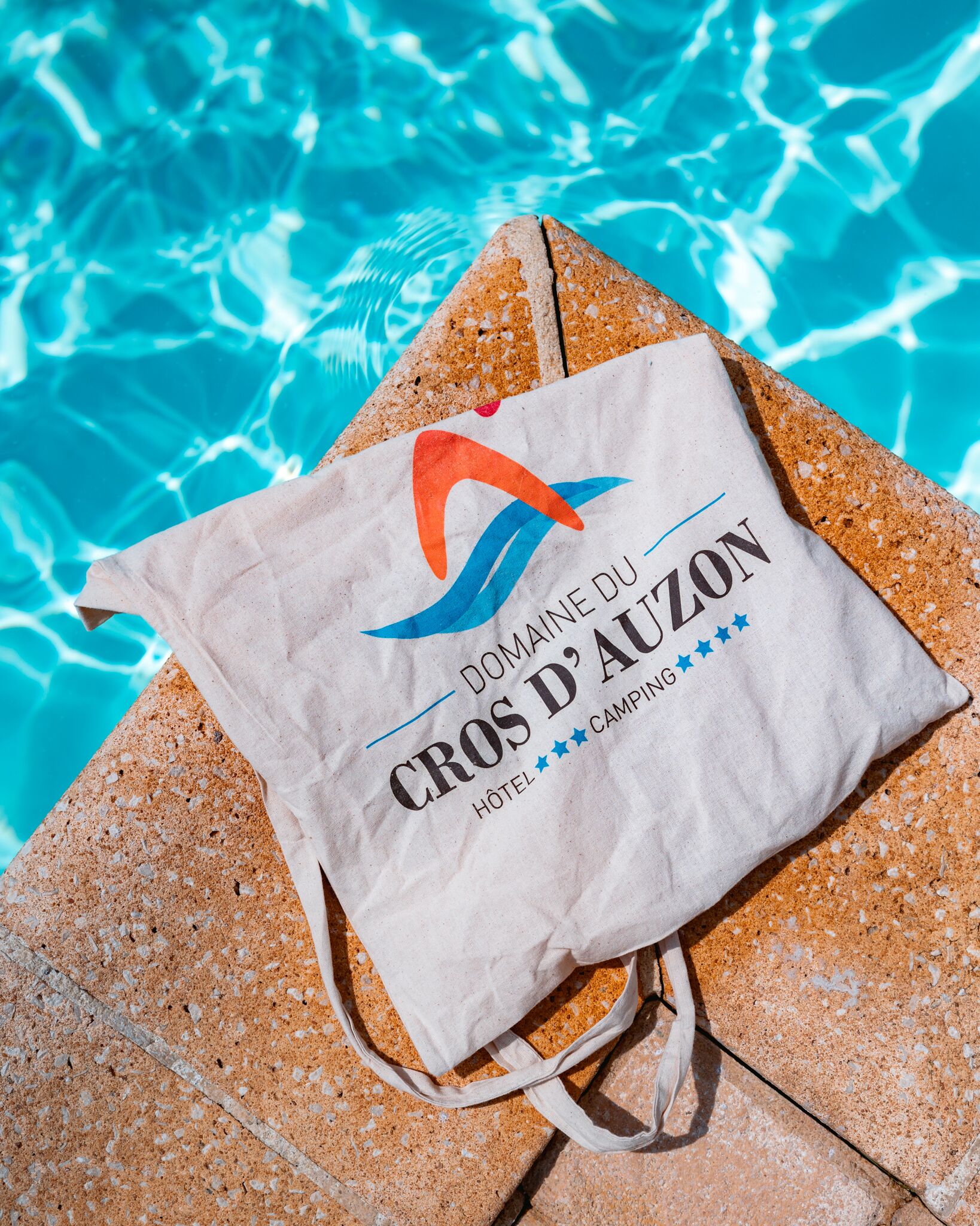 A paper bag with the text "DOMAINE DU CROZ D'AUZON HOTEL CAMPING" placed on a stone surface near a pool.