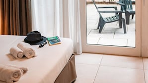 Habitación de hotel con una cama, un juego de toallas, un sombrero y un libro sobre la cama. Hay una puerta corrediza de vidrio que da a un área exterior con sillas y vegetación.