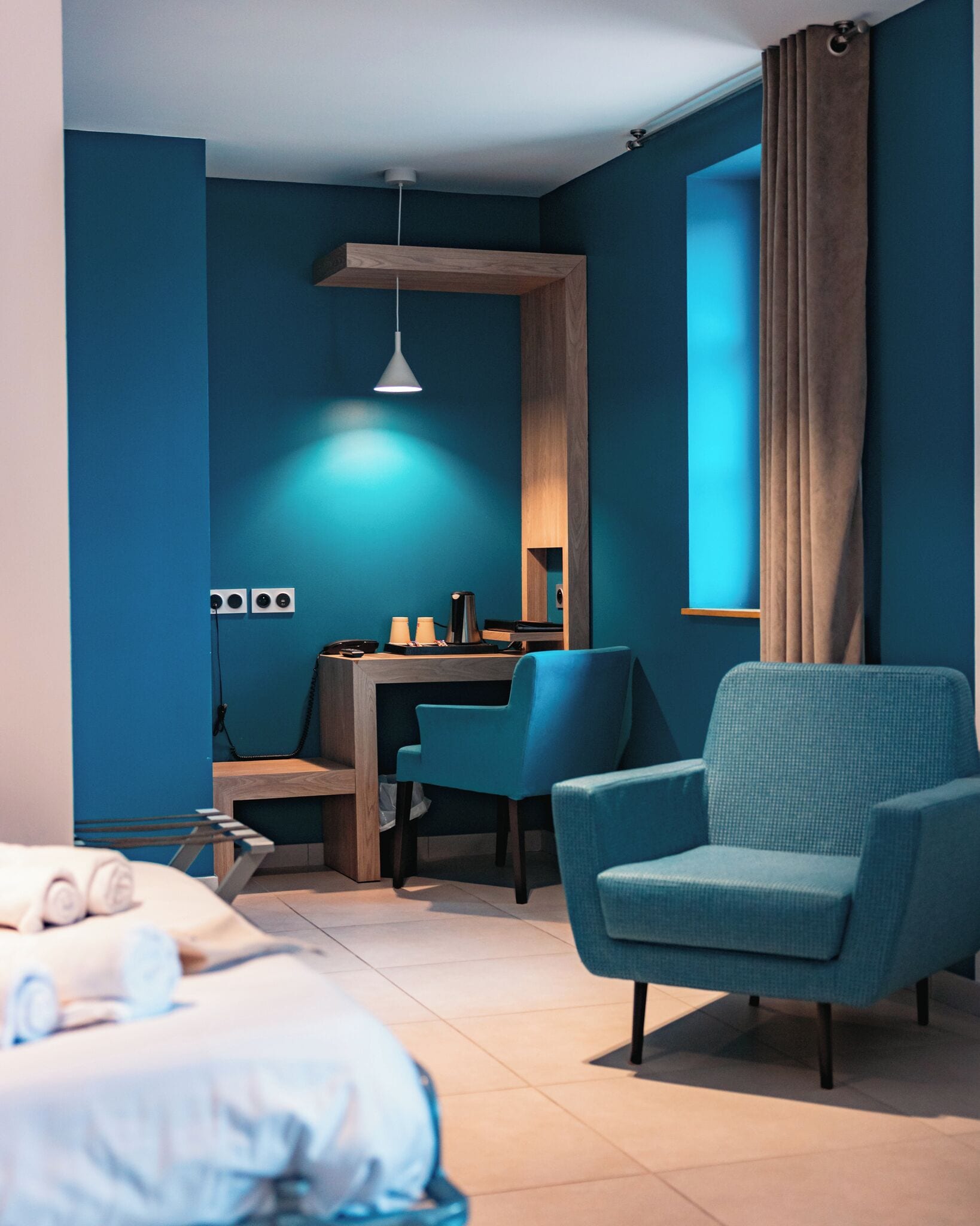 A modern hotel room with a bed, a desk, a chair, a lamp, and a large window with curtains.