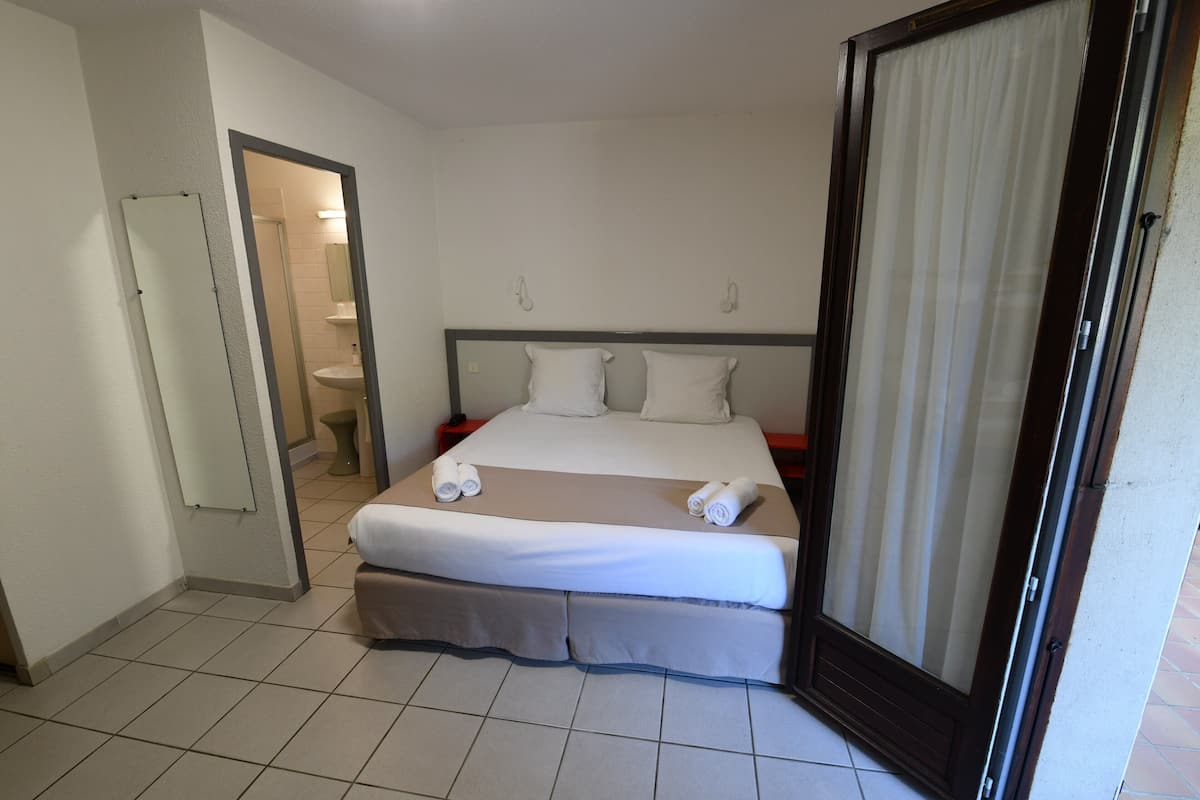 Ein Hotelzimmer mit Bett, einem durchgegangenen Tür sichtbaren Badezimmer und einem mit Vorhängen ausgestatteten Fenster.