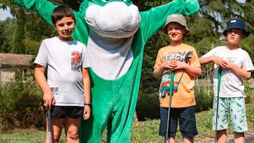 Un personaje con un disfraz verde, rostro de rana y una flor en la cabeza, de pie junto a tres niños que sostienen palos.