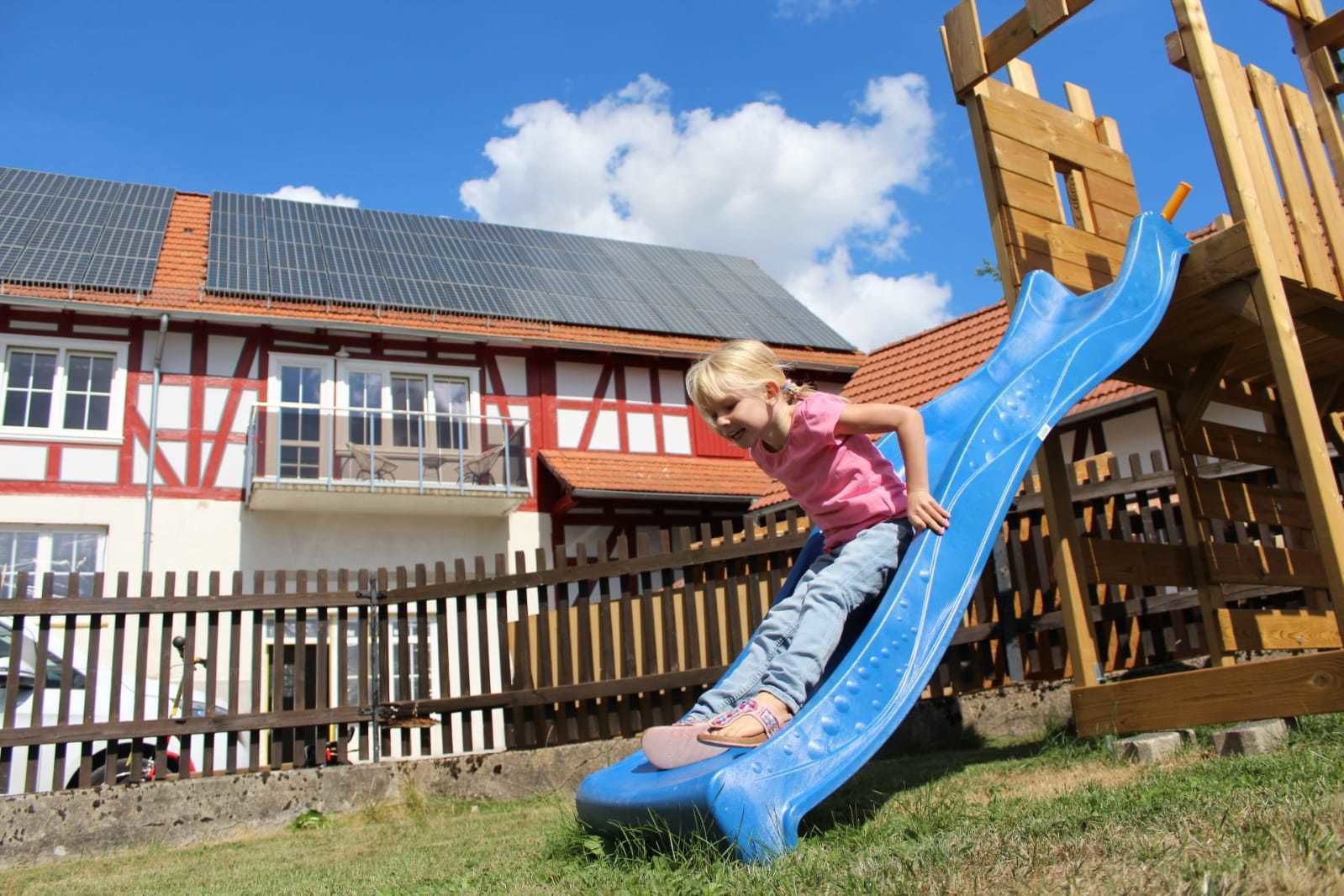 A child on a blue slide in a playground area with a traditional building in the background.