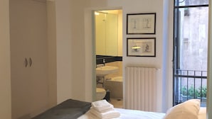 Un dormitorio con una cama, una mesita de noche, un ventanal y un baño visible a través de una puerta abierta.