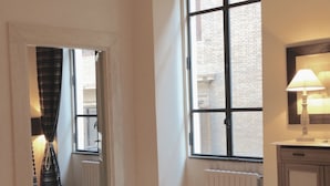 Un pasillo estrecho con un radiador, una ventana que da a un edificio de ladrillo y una lámpara sobre una mesita de noche.