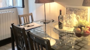 Un comedor con una mesa de vidrio, dos sillas, una lámpara, una botella de vino, copas y un frutero.