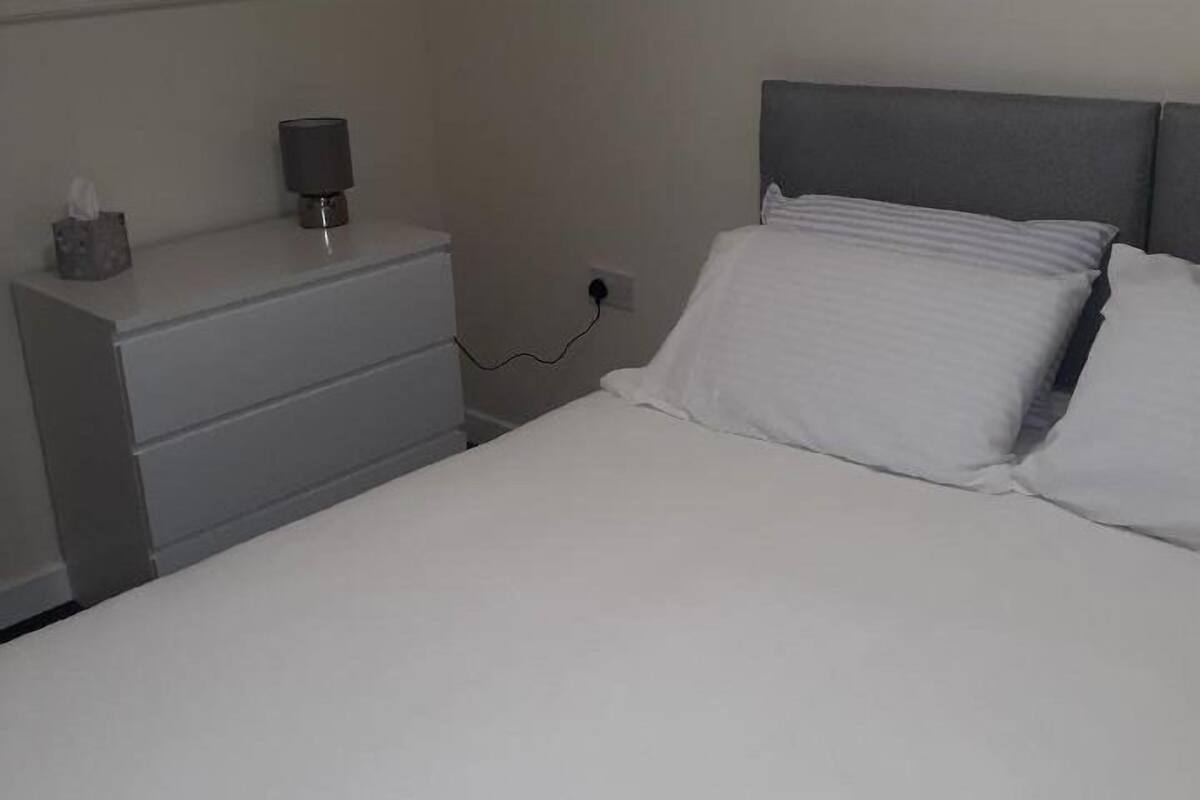 Un lit bien fait, avec du linge de lit blanc et deux oreillers, une table de chevet avec une lampe et une prise électrique sur le mur.