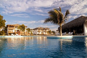 Zona de piscina en un resort con edificios de techo de paja, palmeras y sillas de descanso.