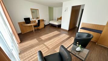 A hotel room with a wooden floor, a desk with a computer, a bed, a chair, and a small table with a vase.