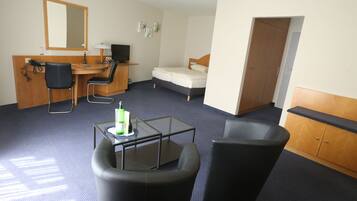 Une chambre d’hôtel équipée d’un bureau, de deux chaises, d’un lit, d’une armoire et d’une petite table sur laquelle se trouve une bouteille.