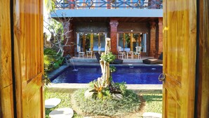 A pool area with a wooden deck, a small waterfall feature, and a view of a building with a balcony. - Pondok DD Canggu (Canggu)