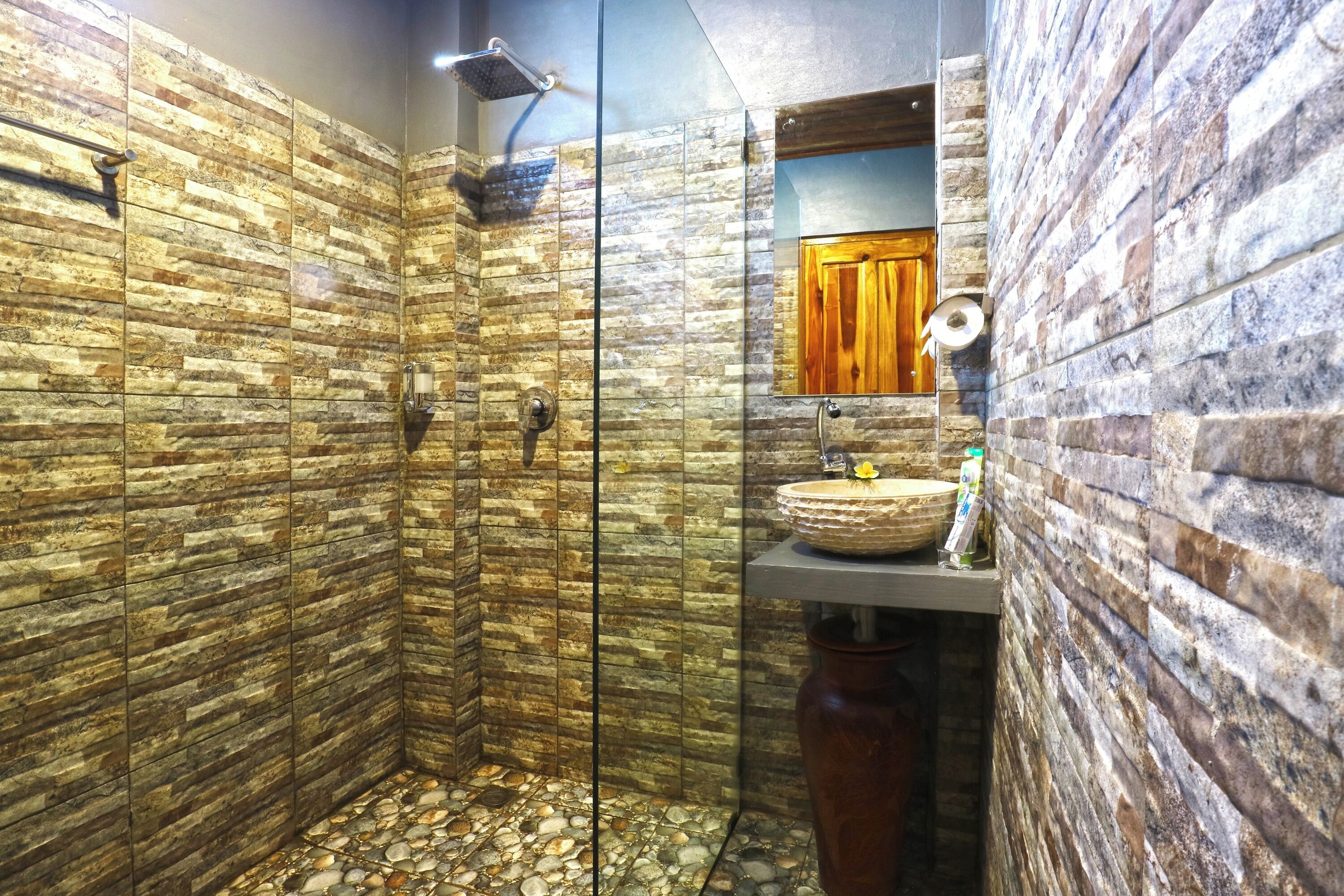 Dutxa amb parets de pedra i porta de vidre, una porta de fusta al fons, i un lavabo amb una bassina rodona.