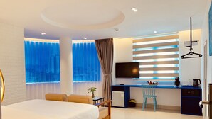 A modern hotel room with a bed, a desk, a chair, a television, and a view of the city through large windows.