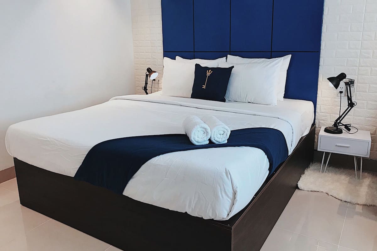 A modern hotel room with a large bed, white bedding, a blue headboard, and a small bedside table with a lamp.